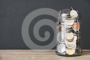 Transparent glass savings jar filled with coins on wooden table with black color wall background, copy space