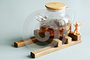 Transparent glass teapot with black flavored tea with added fruit and flower petals on a wooden hot dish stand. Next to a toy