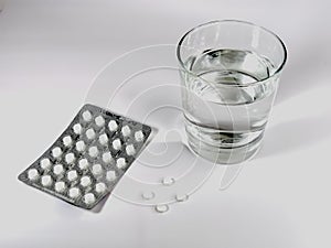 A transparent glass with water, a blister with white tablets and several tablets on a white background