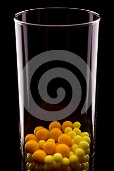 In a transparent glass are yellow and red round tablets. A glass is reflected in the black. Dark background.