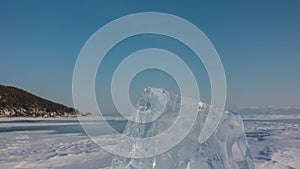 A transparent glittering ice floe stands upright on a frozen and snowy lake.