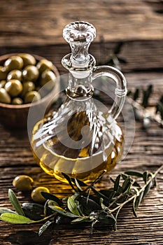 Transparent, golden olive oil is designed with olives in wooden bowl and leaves of olives