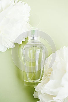 Transparent mockup glass bottle with perfume among fresh white peony flowers on light green pastel color