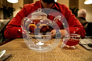 A transparent teapot with fruit tea stands on a table in a cafe in front of a defocused man in a red shirt