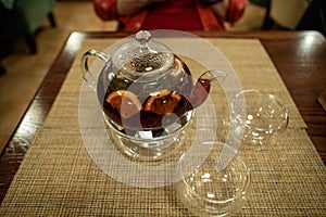 A transparent teapot with fruit tea stands on a table in a cafe in front of a defocused man in a red shirt