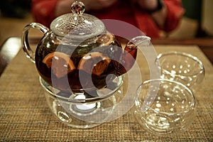 A transparent teapot with fruit tea stands on a table in a cafe in front of a defocused man in a red shirt