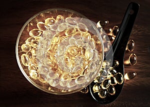 Transparent yellow pills in bowl on wood background, capsules cod liver oil, omega 3 in ceramic black spoon.