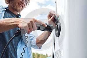 Transportation. Young man traveling by electric car stop at chraging station plugging in charger close-up smiling