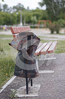 The trash can is damaged, broken in the Park near the bench. Vandalism