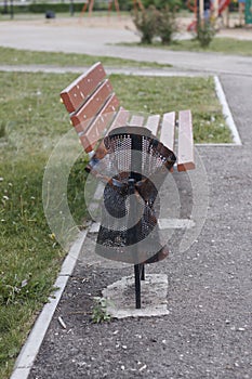 The trash can is damaged, broken in the Park near the bench. Vandalism