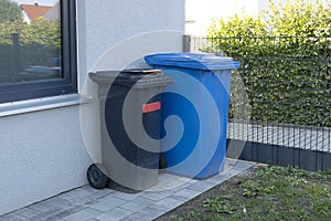 Trash cans of black and blue on wheels stand near the wall. Dirty mobile trash bins and household waste. Sorting garbage into