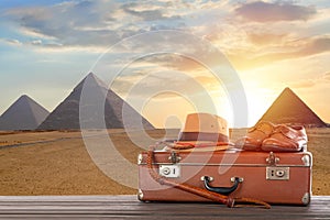 Travel, archeology and adventure concept. Vintage brown suitcase with fedora hat, bullwhip and shoes against Great pyramids in