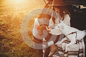 Travel and autumn road trip with pet. Stylish hipster woman in sweater holding phone and sitting with cute dog in car trunk in