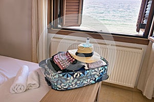 A travel bag with female clothes, straw hat, mask, hand sanitiser over a hotel room.