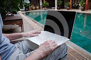 Travel blogger sitting at swiming pool writing article on white laptop