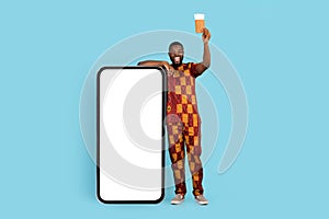 Travel Bookings Online. African Guy In Traditional Costume Standing Near Blank Smartphone