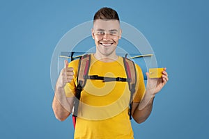Travel budget. Attractive young man with tourist gears gesturing thumb up and showing credit card on blue background