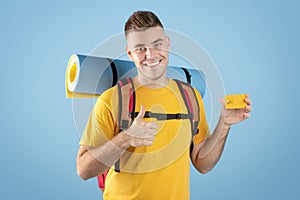 Travel budget. Cool millennial guy with backpack and credit card showing thumb up gesture over blue studio background