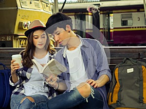 Travel concept.couple of love reading book wating train.Asian beautiful girl wearing hat holding coffee cup and smart man holding