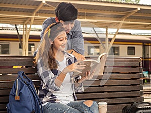 Travel concept.couple of love reading book wating train.Asian beautiful girl wearing hat holding coffee cup and smart man holding