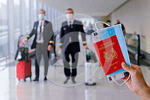 Travel concept: hand holding passport, mask, boarding pass in airport. Two pilots walking with luggage in background