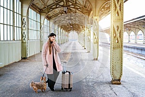 Travel concept. At station, young tourist with dog goes and drags suitcase