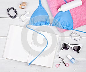 blue swimsuit, book, pink towel, cosmetics makeup, bijou and essentials on white wooden desk