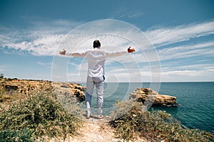 Travel concept. Young tourist man standing on the edge of reef enjoying ocean or sea view of turquoise water with raised hands