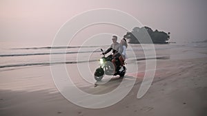 Travel couple riding motorbike on ocean sandy beach in tropical paradise at sunset. Man with girlfriend driving