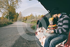 Travel in fall season. Father and his little child sitting inside car trunk near rural road and looking at map. Happy family