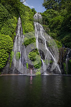 Travel lifestyle. Young traveler woman standing in front of waterfall in tropical forest. Excited woman raising arms. Freedom