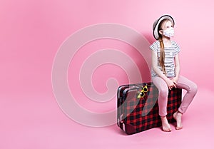 Travel: A little girl in a straw hat and a medical mask is sitting on a big red suitcase and waiting for travel.