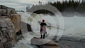 Travel man standing in front of waterfall in norway forest at early morning. Dressed in a red hat.