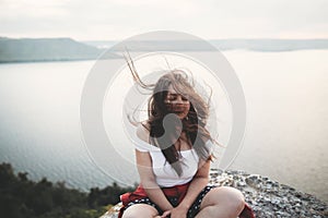 Travel and Meditation. Hipster girl with windy hair sitting and meditating on top of rock mountain with beautiful view on river.