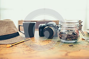 Travel money savings in a glass jar, camera, coffee cup, straw hat and pen on world map. Travel budget concept