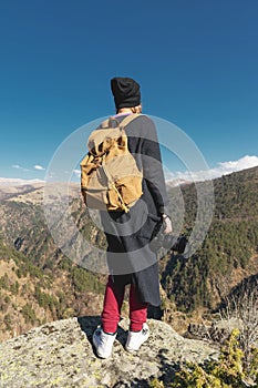 A travel photographer girl hipster in a hat and with a backpack standing on a rock and looking at the hills.