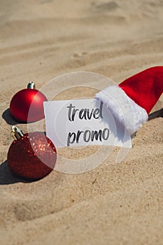 TRAVEL PROMO text on paper greeting card on background sandy beach sun coast. Christmas balls Santa hat New Year New Me