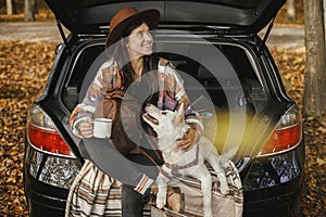 Travel and road trip with pet. Stylish happy woman traveller with cup and backpack sitting with cute dog in car trunk in sunny