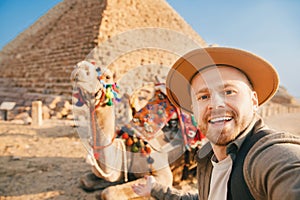 Travel selfie photo tourist man in hat with camel background pyramid of Egyptian Giza, sunset Cairo, Egypt