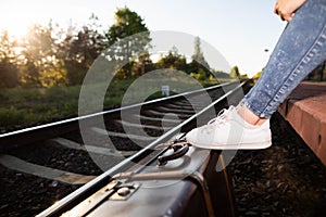 The old suitcase stands right next to the railway tracks on the platform and on it are the feet in the sports shoes of