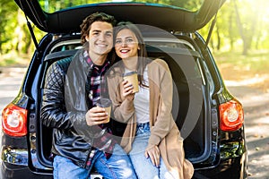 Travel, summer vacation, road trip, leisure and people concept. Happy couple drinking coffee from disposable cups sitting on trunk