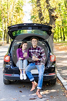 Travel, summer vacation, road trip, leisure and people concept. Happy couple drinking coffee from disposable cups sitting on trunk