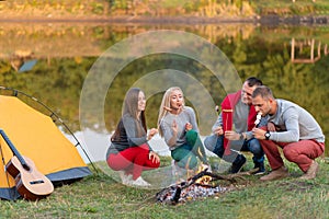 Travel, tourism, hike, picnic and people concept - group of happy friends frying sausages on campfire near lake