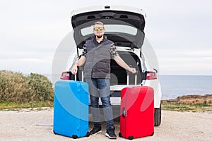 Travel, tourism and trip concept - young man sitting in open car trunk with two suitcases and smiling