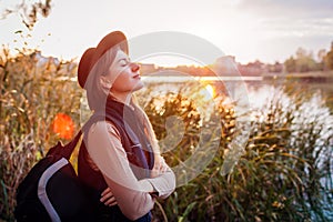 Traveler with backpack relaxing by autumn river at sunset. Young woman breathing deep feeling happy and free