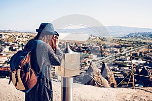 A traveler with a backpack on at viewpoint looks through binoculars at a beautiful view of the town of Goreme in