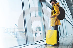 Traveler in bright jacket with yellow suitcase backpack at airport on background large window blue sky, passenger waiting flight