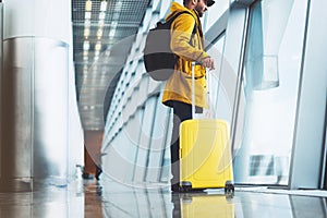 Traveler in bright jacket with yellow suitcase backpack at airport on background large window blue sky, passenger waiting flight