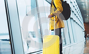 Traveler in bright jacket with yellow suitcase backpack at airport on background large window blue sky, passenger waiting flight