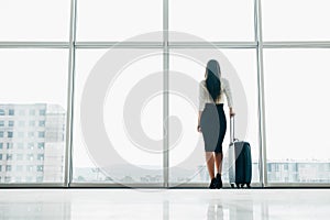 Traveler businesswoman waiting for delayed flight at airport lounge standing with luggage watching tarmac at airport window. Woman